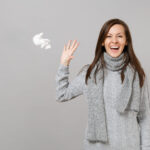 Happy young woman in gray sweater, scarf recovering and throwing paper napkin isolated on grey wall background. Healthy lifestyle, ill sick disease treatment, cold season concept. Mock up copy space.