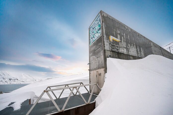 Snow-covered Svalbard Global Seed Vault building with a metal ramp leading to the door against a pale blue sky.
