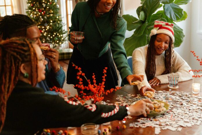 Friends sit around a table, making holiday cookies with a Christmas tree in the background.