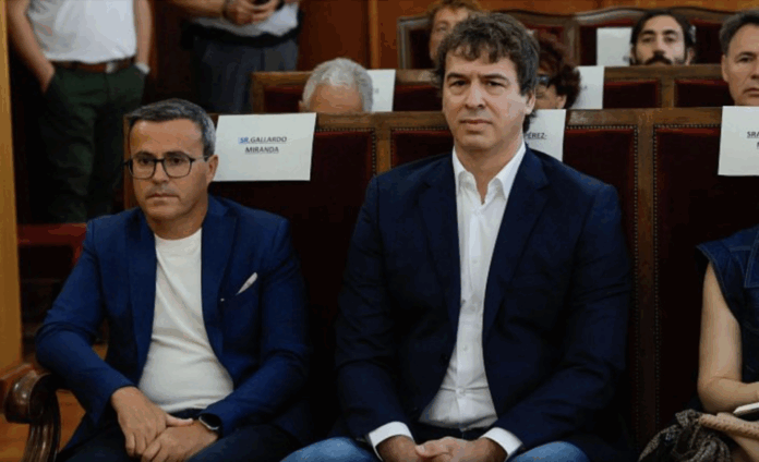 Two men in blue blazers sit in a formal auditorium, facing forward during an event.
