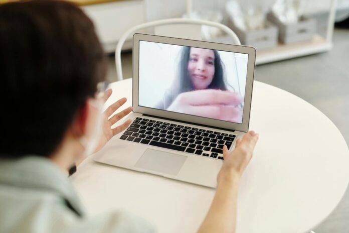Person video calling on a laptop at a white table, screen shows a smiling woman on the call, blurred foreground of the caller's head and hands