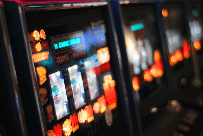 Row of brightly lit slot machines in a casino with glowing screens and red button lights along the front panel, blurred background to convey atmosphere.