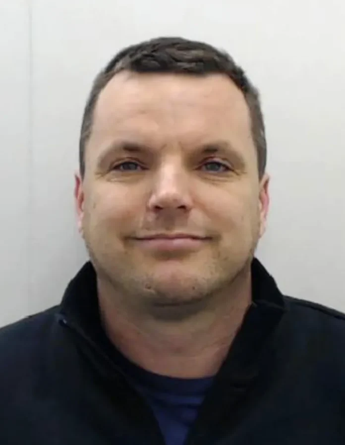 Head-and-shoulders portrait of a man with short dark hair, wearing a black jacket and blue shirt, smiling softly against a light gray background.