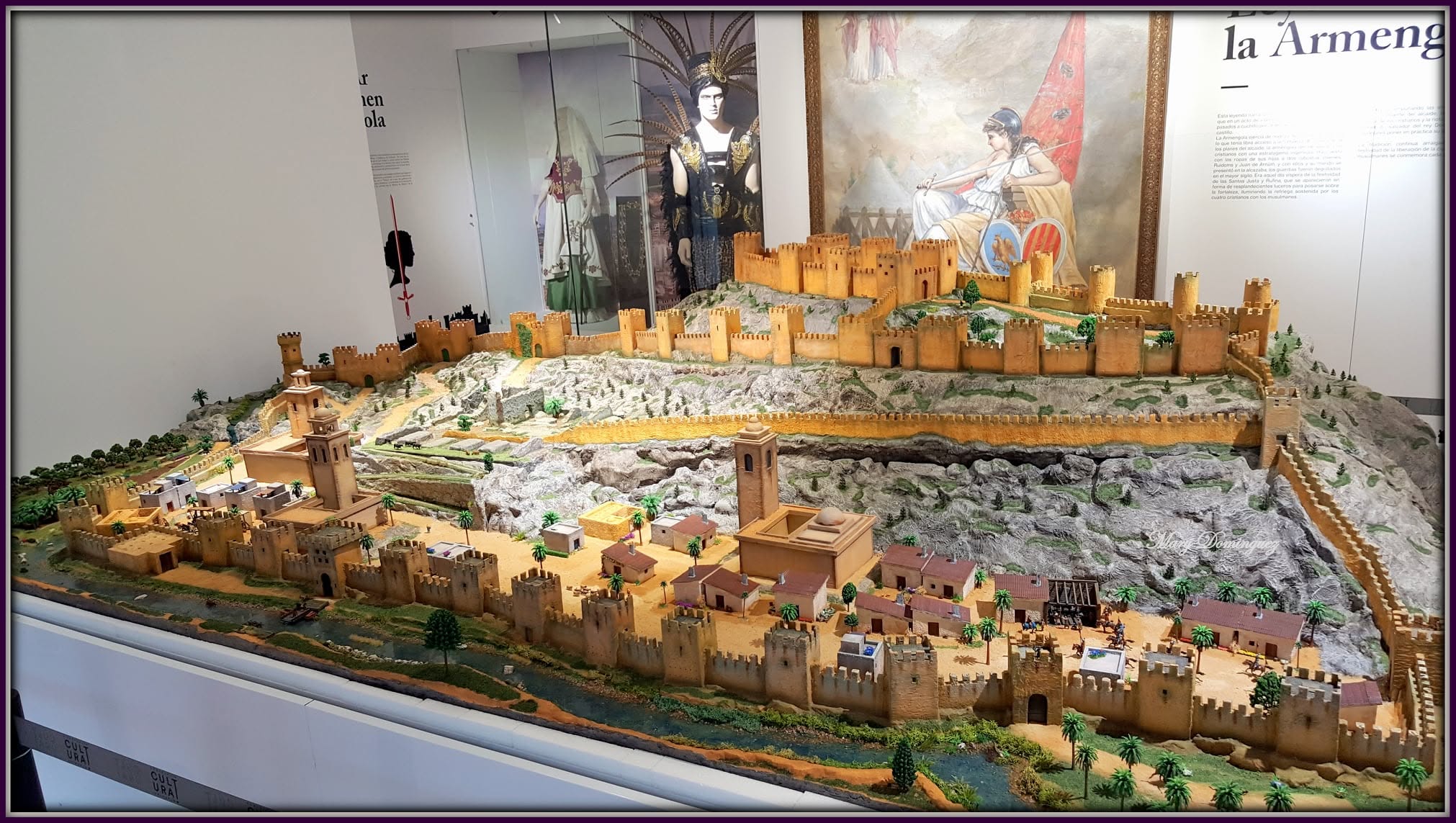 Scale model of a walled fortress on a rocky hill, with towers, gates, and a surrounding village displayed in a museum diorama