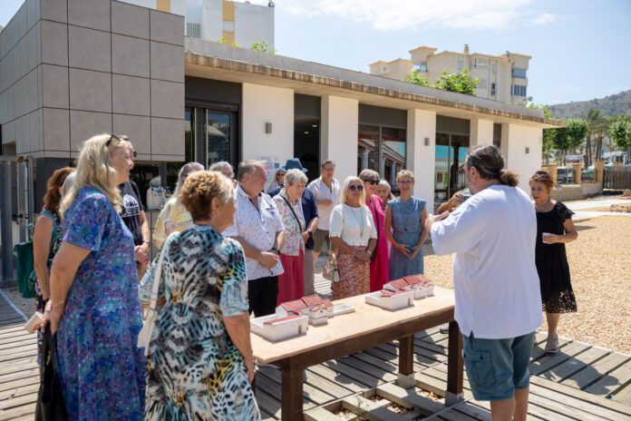 Group of adults in colorful summer clothing gathered around a table with scale-model houses outside a modern building.