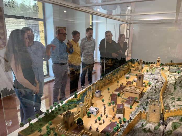Group of adults examining a detailed desert village diorama inside a glass display case.