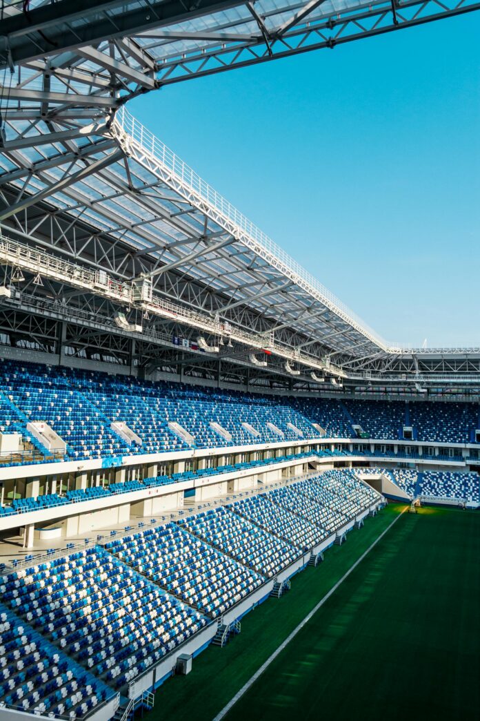 Interior of a large stadium with blue seating, green turf, and white steel framework overhead under a clear blue sky.