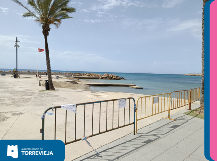 Coastal promenade cordoned off with barriers, a palm tree, and calm blue sea in the background; Torrevieja branding visible in the corner.