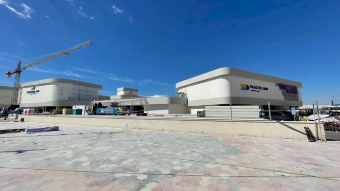 White curved shopping plaza under construction, crane to the left, blue sky above.