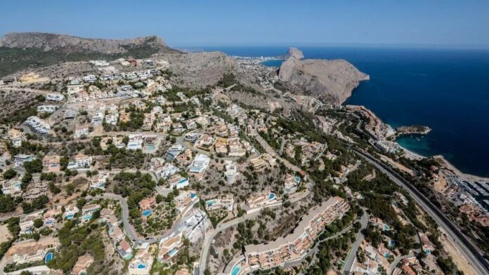 Aerial view of a coastal hillside town with winding roads and clustered white and beige houses overlooking a deep blue sea.