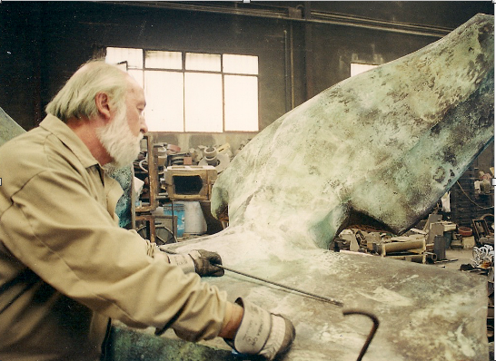 Older man with gloves smoothing a large green curved metal panel in a workshop, using a tool on it.