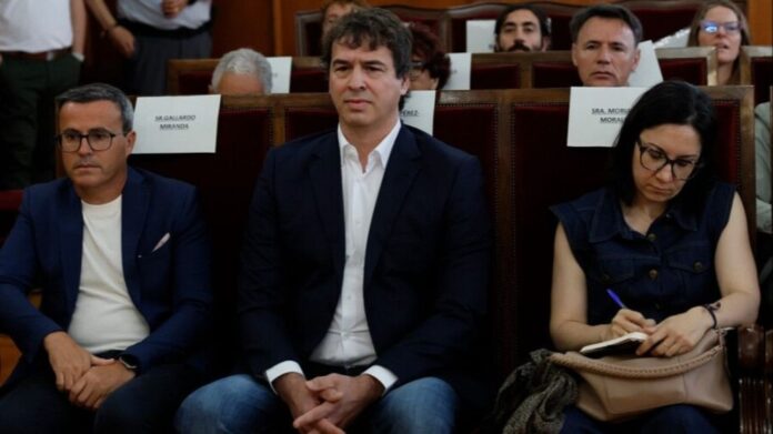 Three people sit in a formal meeting hall; center man in a dark blazer and white shirt, left man with glasses, right woman writes in a notebook.