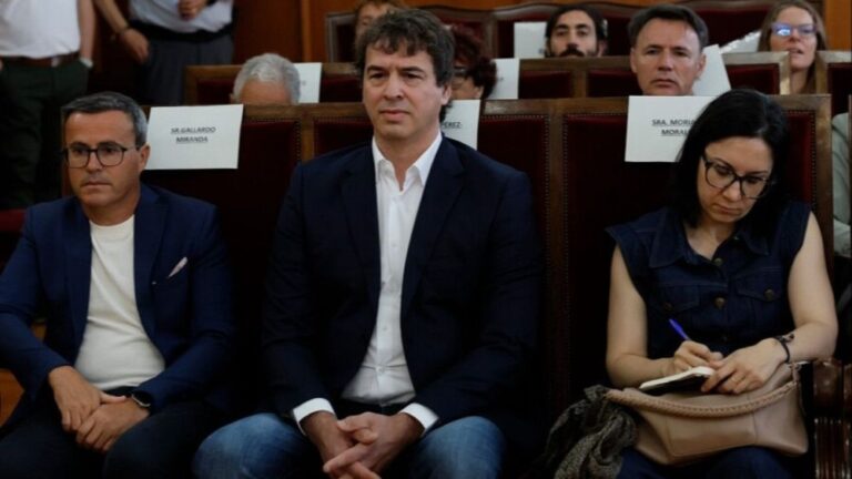 Three people sit in a formal meeting hall; center man in a dark blazer and white shirt, left man with glasses, right woman writes in a notebook.