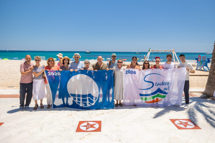 Group of about 14 adults on a sunny beach holding banners that read 2026 and a logo, with the sea in the background.