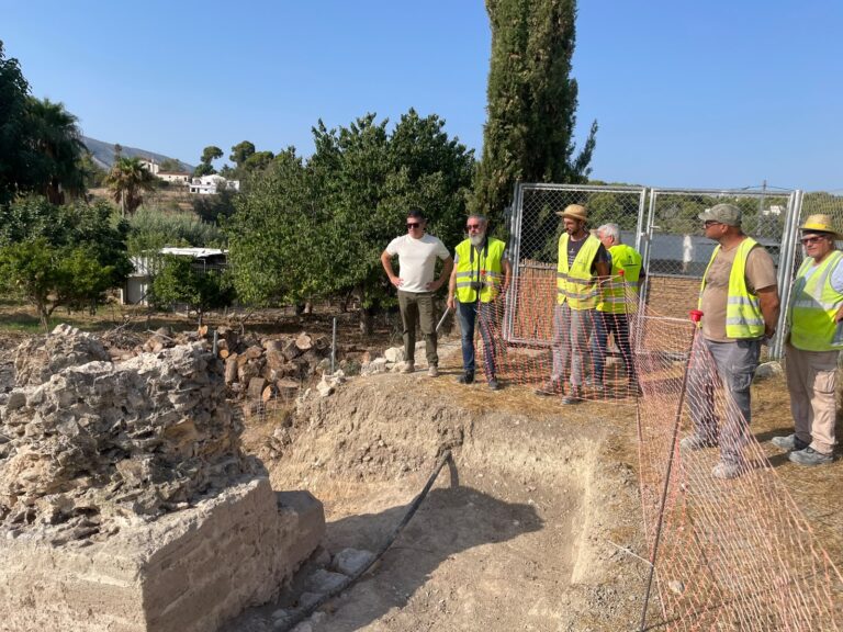 Group of workers in yellow safety vests standing by an orange construction barrier at a rocky excavation site on a sunny day.