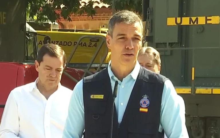Man in a light blue shirt and black vest speaks at a microphone during an outdoor briefing, with industrial machinery in the background.