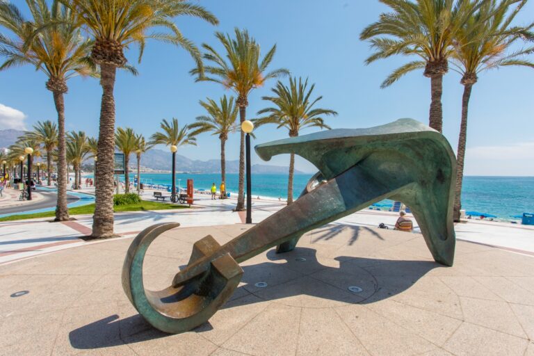 Bronze abstract anchor sculpture on a seaside promenade with palm trees and blue sea.