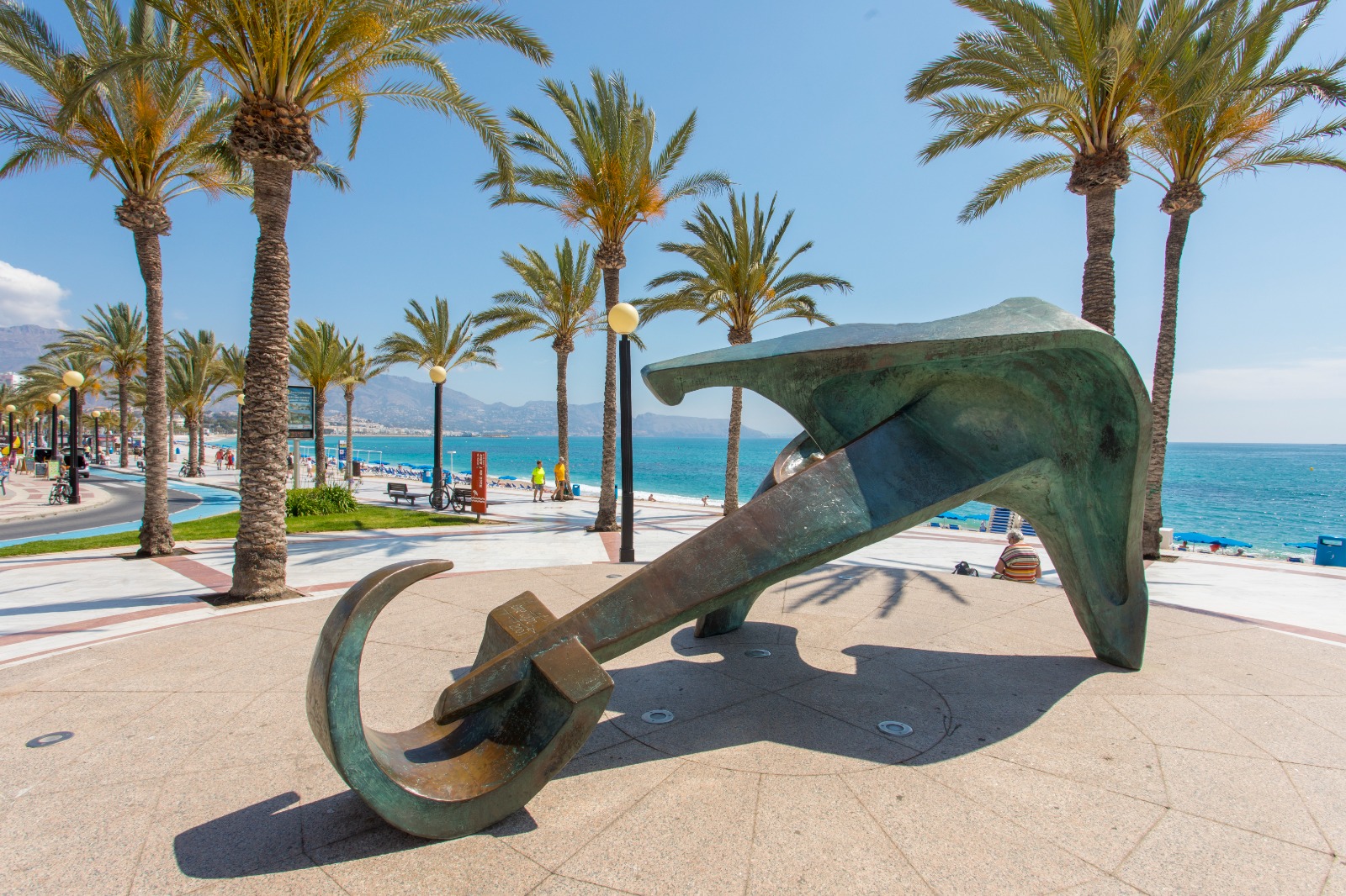 Bronze abstract anchor sculpture on a seaside promenade with palm trees and blue sea.