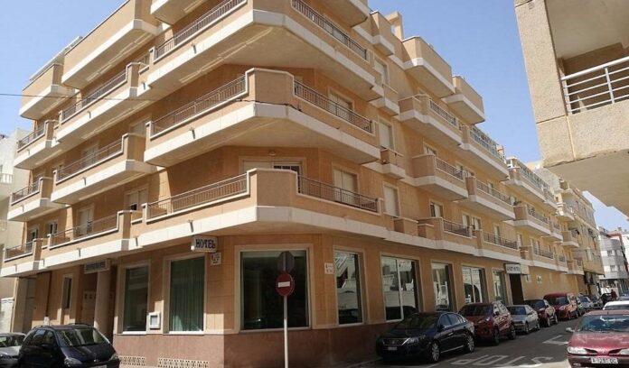 Corner street building with beige balconies and storefronts on ground floor, cars parked along curb.