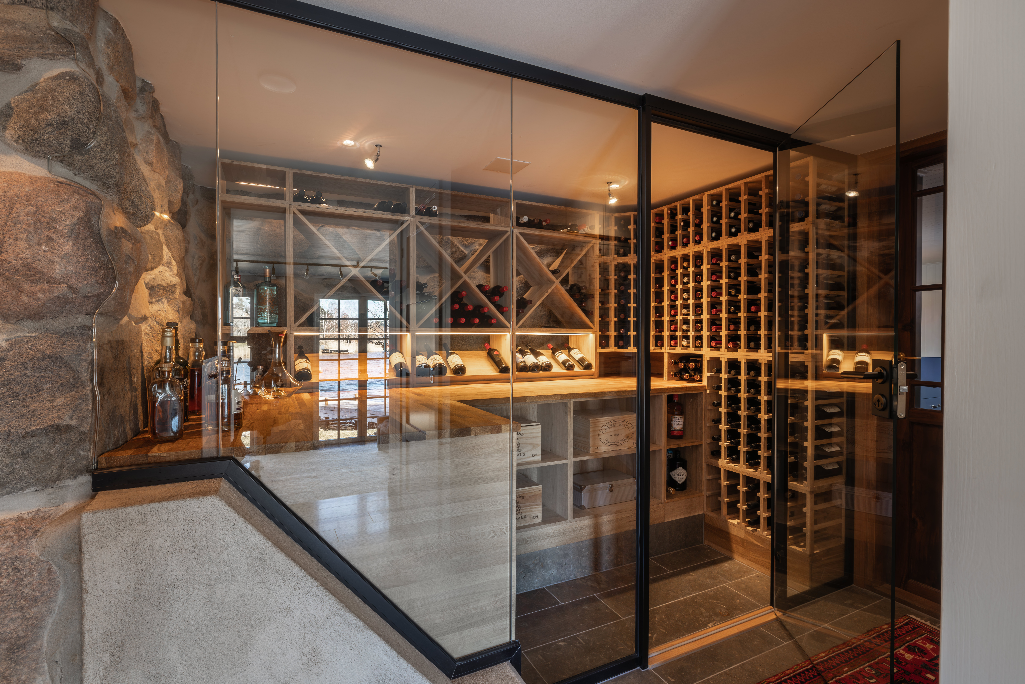 Wine cellar with glass doors, wooden racks filled with wine bottles and a stone wall on the left, warm lighting above.
