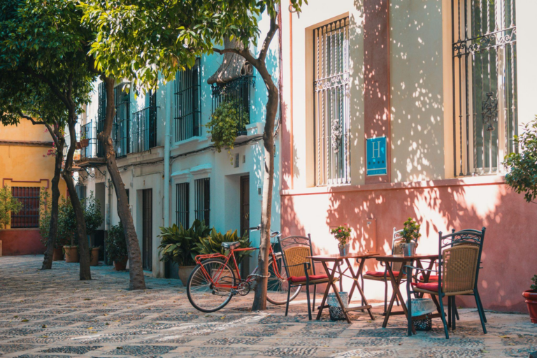 Cobbled street lined with pastel houses, leafy trees, and an outdoor cafe with tables and chairs.