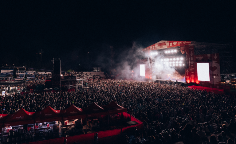 Nighttime outdoor concert with a colossal crowd facing a brightly lit stage and smoke effects behind it.