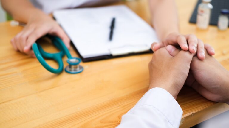 Medical professional and patient hold hands across a wooden table, with a stethoscope and clipboard nearby.
