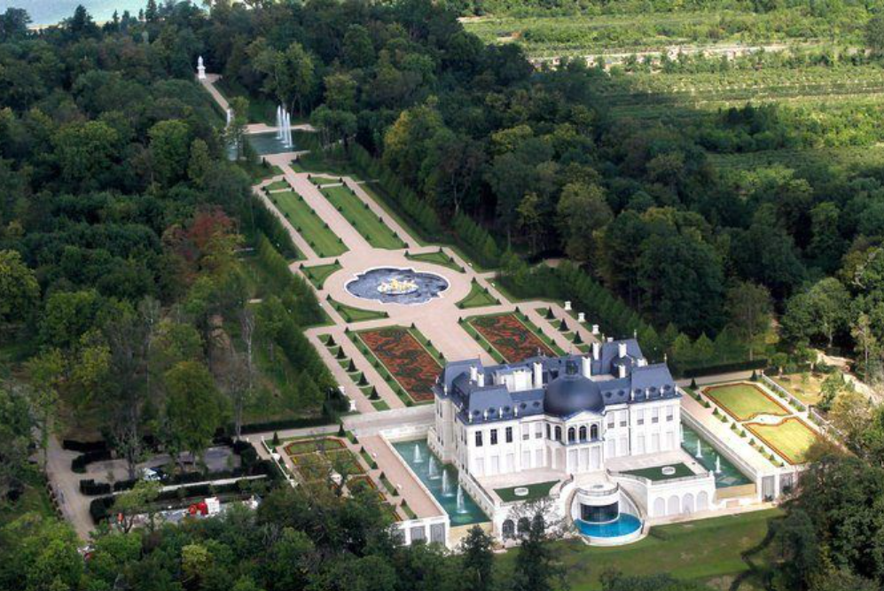 Aerial view of a white European-style palace with a blue dome, surrounded by formal gardens and reflecting pools.