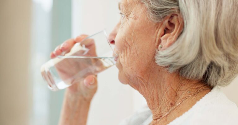 Older woman drinks water from a glass, shown in profile.