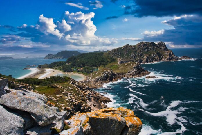 Rugged coastline with rocky cliffs, a sandy beach, and blue sea under a cloudy sky.