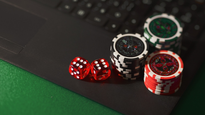 Two red dice with white pips sit beside stacked poker chips on a dark desk near a laptop keyboard and green felt Casino surface