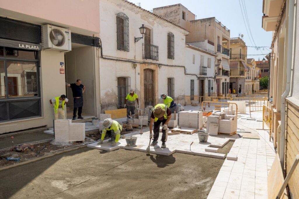 Workers in high-visibility vests lay white tiles on a sidewalk beside a storefront labeled 'LA PLAZA'.