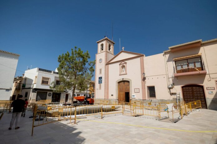 Church with a bell tower in a plaza under construction, surrounded by yellow barricades and a tree against a clear blue sky.