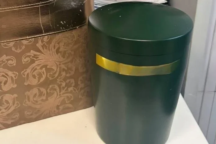 Dark green cylindrical trash can with a gold band around the lid, placed on a white surface beside a brown patterned box in a room corner.