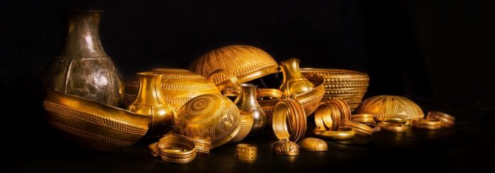 Collection of ornate gold jewelry and vessels arranged on a dark surface, catching warm light.