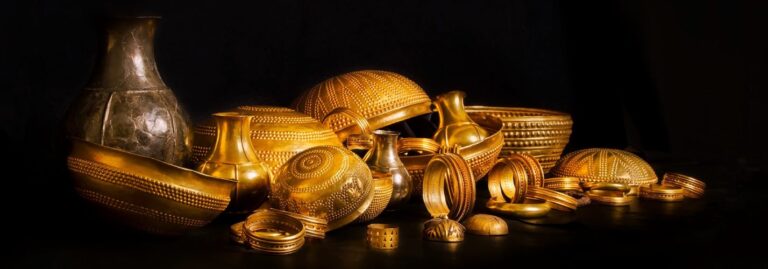 Collection of ornate gold jewelry and vessels arranged on a dark surface, catching warm light.